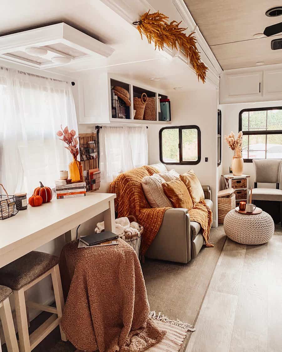 Cozy RV interior with a sofa, orange blankets, pillows, and autumn decor; white table, stools, and pouf complete the inviting space