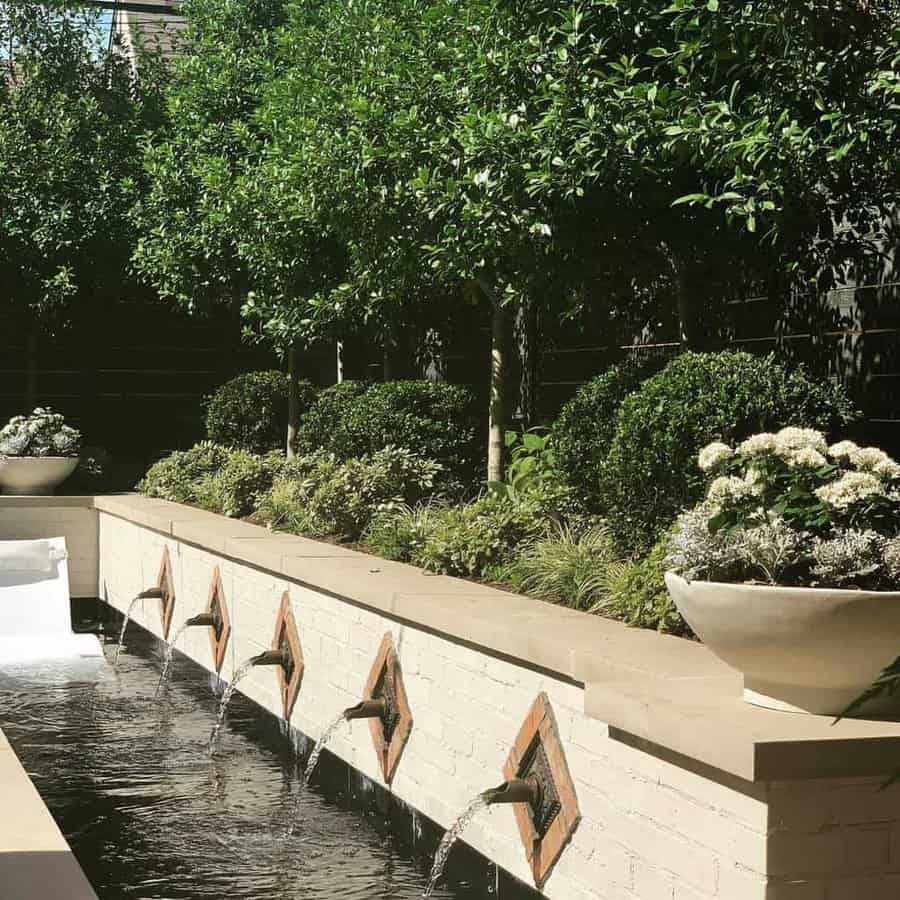 Garden with a brick wall fountain, water spilling from spouts into a narrow pool, surrounded by lush greenery and potted plants