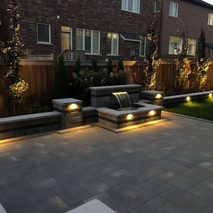 Modern backyard with illuminated water feature, stone tiles, and ambient lighting, fenced garden and townhouse in the background