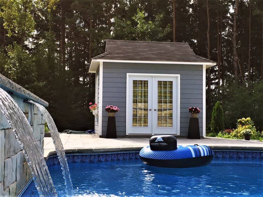 A backyard pool with a waterfall feature, an inflatable tube, and a gray shed with double doors surrounded by trees and plants