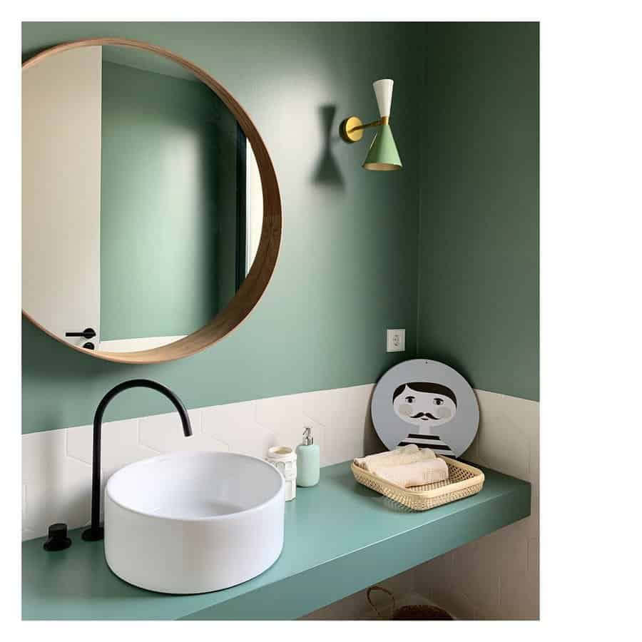 Chic bathroom with soft green walls, minimalist white hexagon tile backsplash, round vessel sink, matte black faucet, and playful accents.