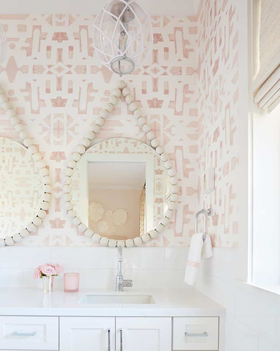 Charming bathroom with glossy white tile backsplash, playful blush patterned wallpaper, round beaded mirrors, and soft pastel decor.