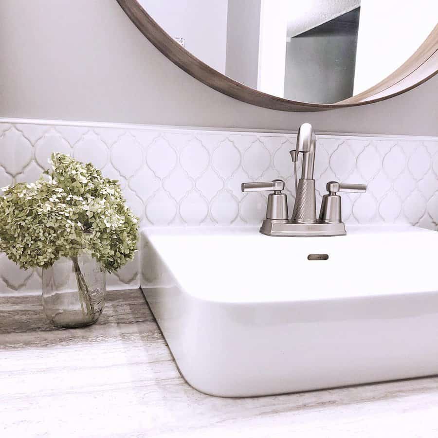 Elegant bathroom with white arabesque tile backsplash, modern brushed nickel faucet, round mirror, and a soft wood countertop.