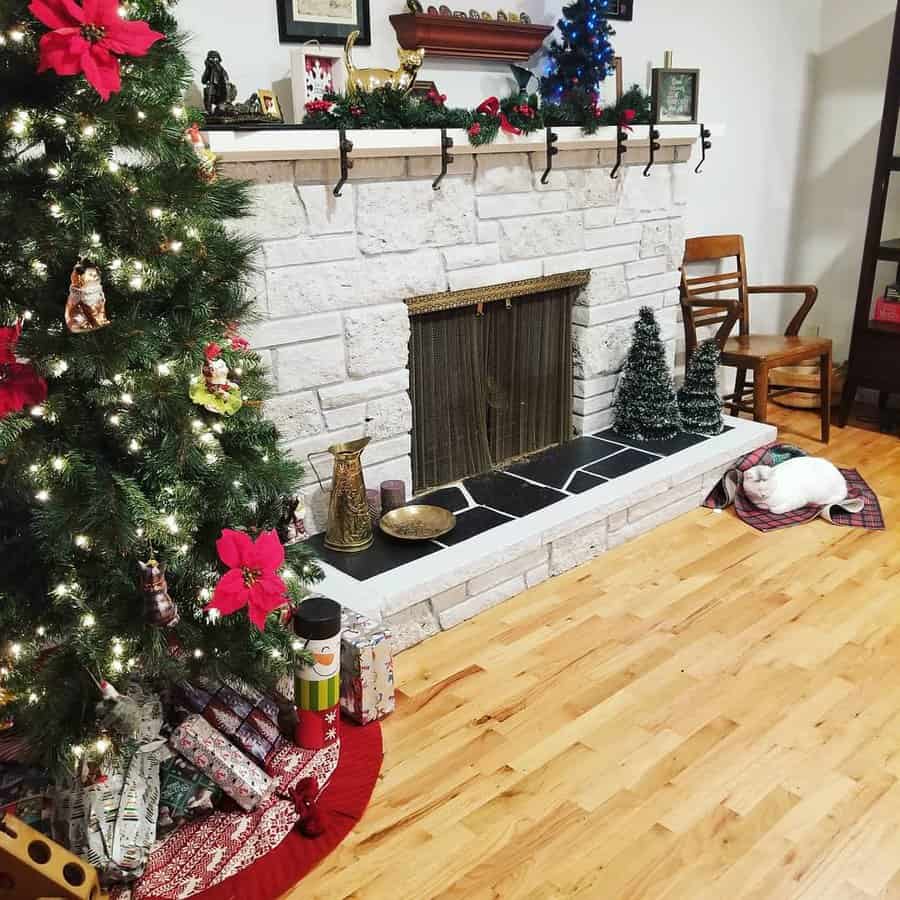 A festive stone fireplace decorated with holiday garlands, stockings, and two small Christmas trees, with wrapped gifts beneath the tree, creating a warm holiday ambiance