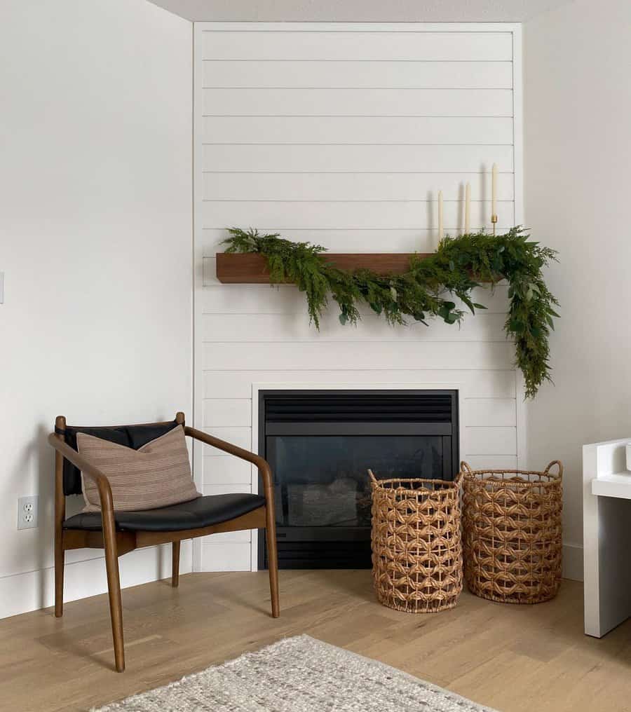 A minimalist white fireplace with a wooden mantel, decorated with a greenery garland and candles, paired with a modern chair and woven baskets for a clean, cozy look