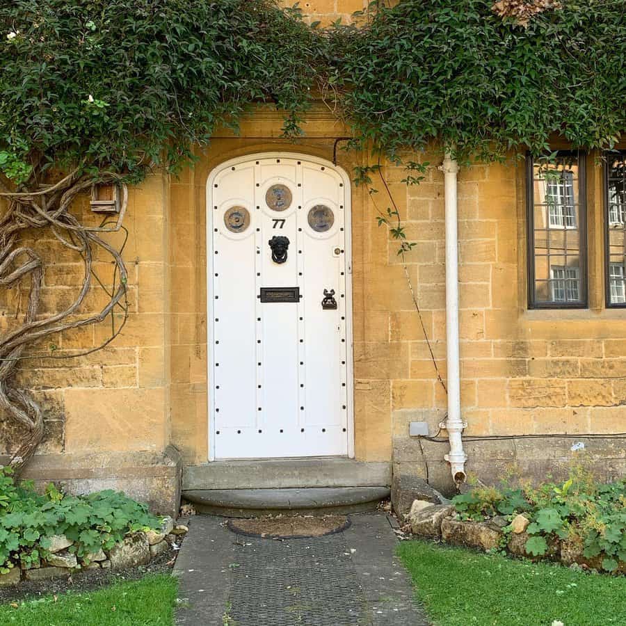 White arched door with circular windows, surrounded by ivy on a yellow brick house, black knocker and number "77" on the door