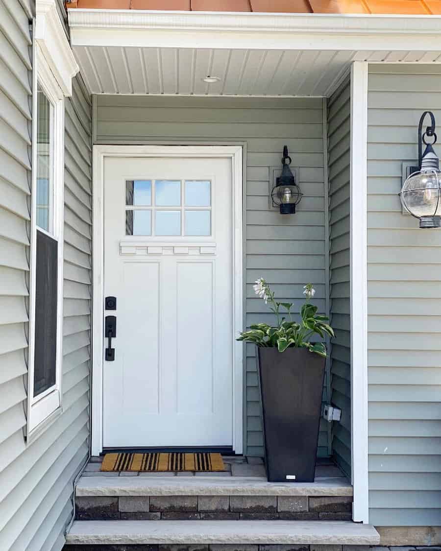 White front door with black hardware, flanked by green siding; tall black planter with white flowers; two lanterns on either side