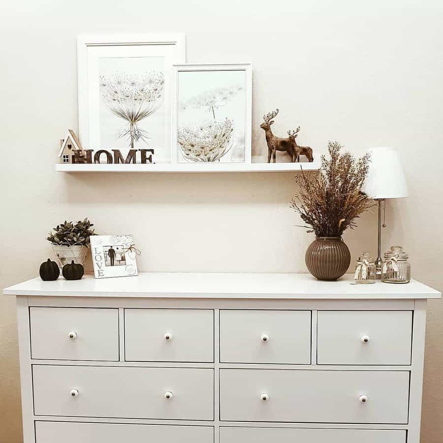 Minimalist white dresser with matching knobs, styled with neutral decor, framed botanical prints, and a floating shelf with rustic accents.