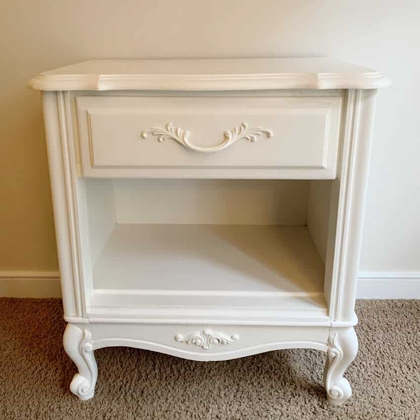 Elegant white-painted nightstand with ornate detailing, curved legs, and a single drawer, blending classic French-inspired design with vintage charm.