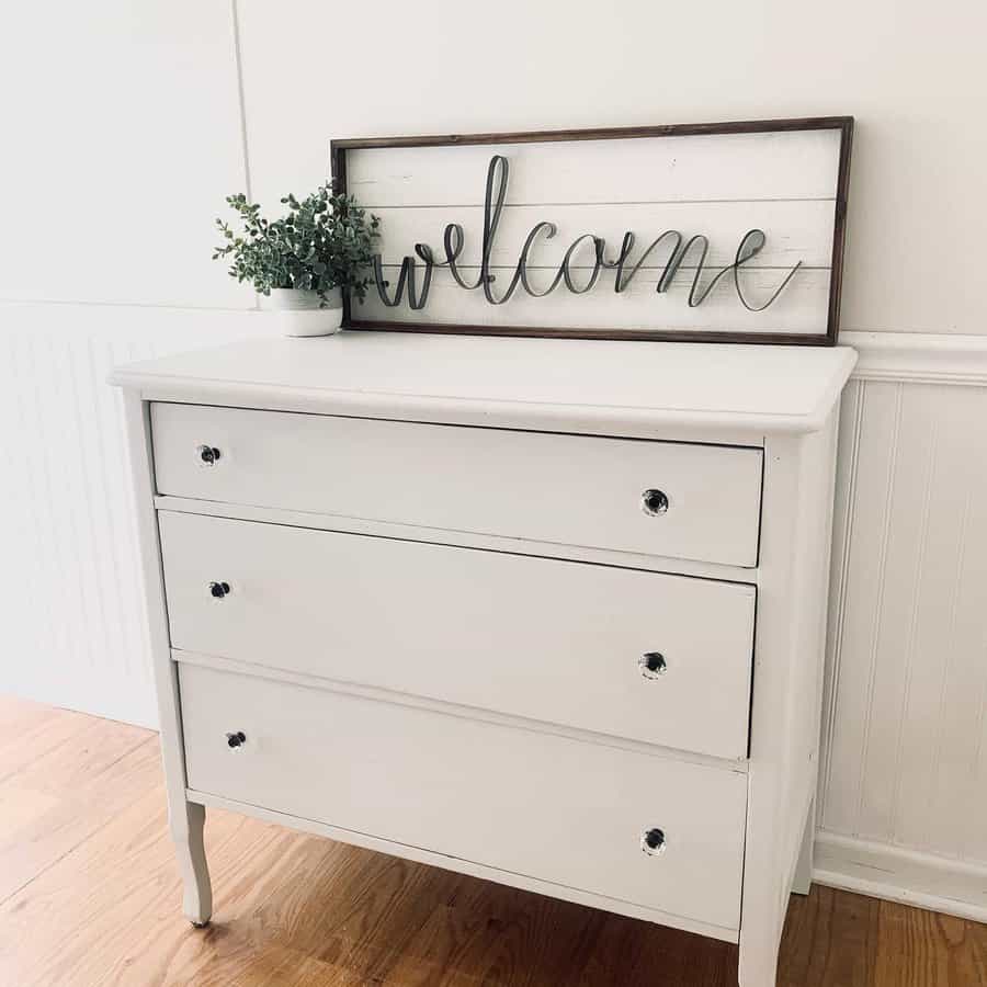 Simple white-painted dresser with glass knobs, styled with a farmhouse-style 'Welcome' sign and a small potted plant on a wood floor.