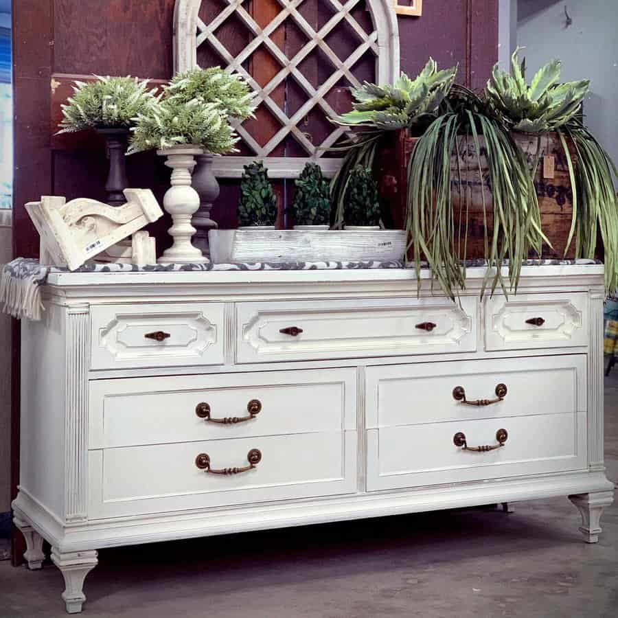 Vintage white-painted dresser with ornate brass handles, styled with lush greenery, decorative accents, and a rustic farmhouse backdrop.