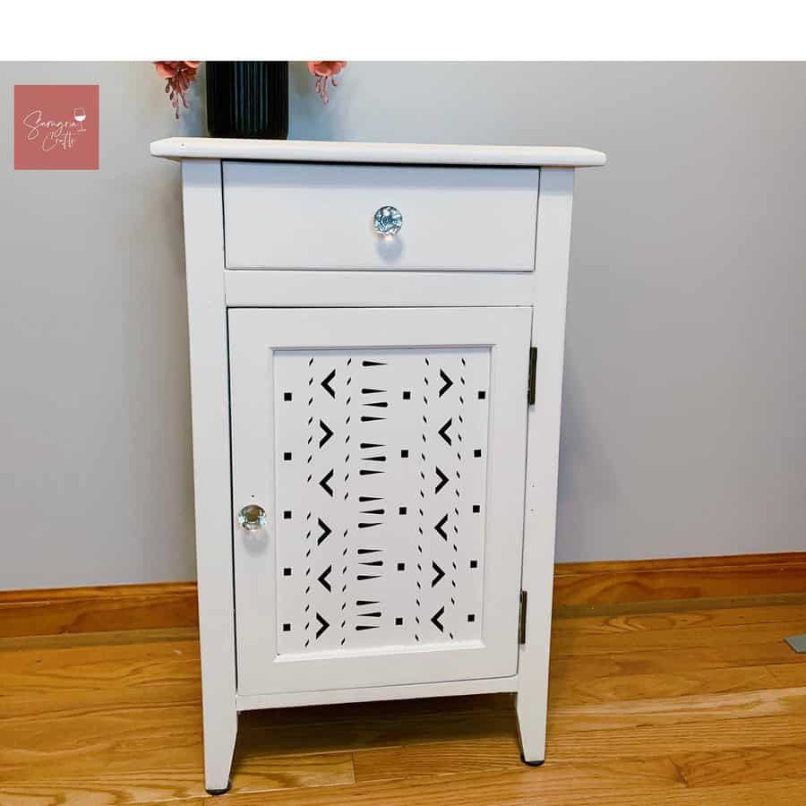 White-painted farmhouse-style cabinet with a decorative cutout door panel and glass knobs, set on a warm wooden floor.