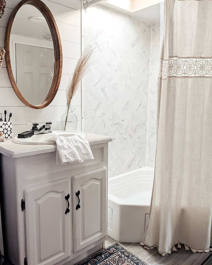 A cozy RV bathroom with a round mirror, white vanity, faucet, and a shower with a lace-trimmed curtain, decorated with a vase of reeds.