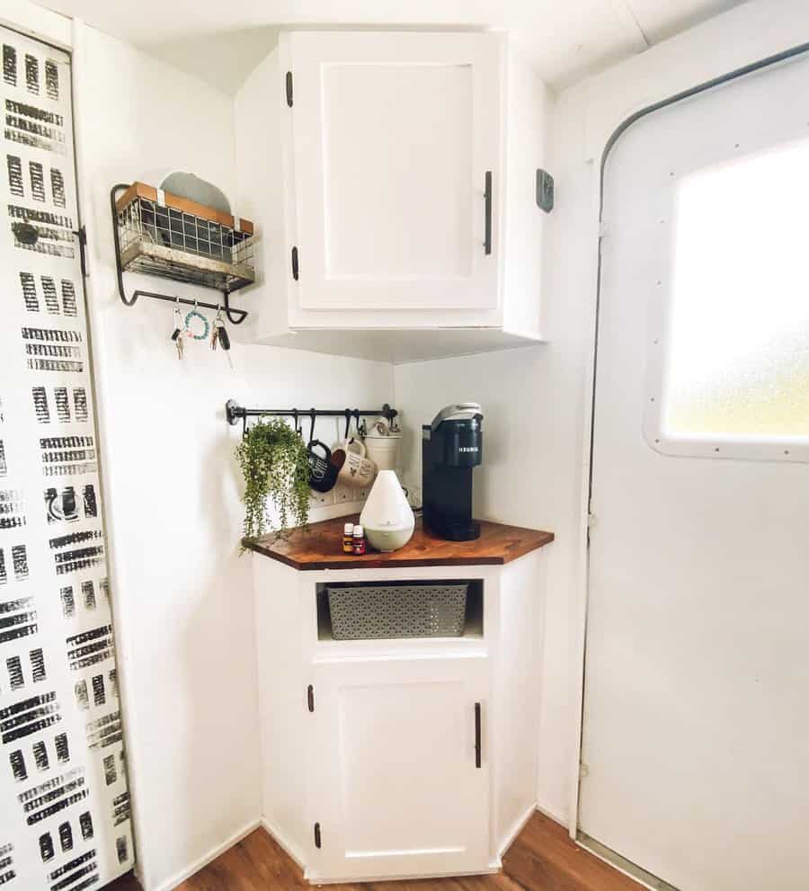 Small white RV nook with wooden countertop, coffee maker, and potted plant; wall shelf and hooks hold kitchen items; door to the right.