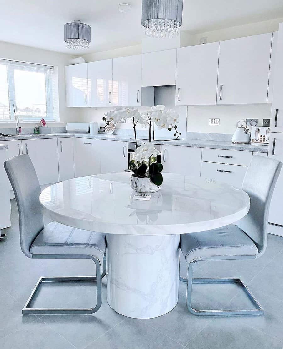 Modern white kitchen with a round marble table, two gray chairs, and an orchid centerpiece, featuring white cabinets and gray floor tiles