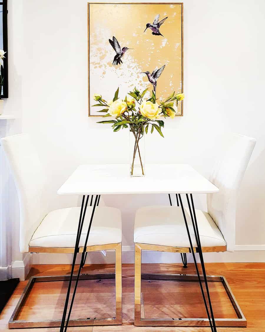 Small white dining table with two chairs, a vase of yellow flowers, and a hummingbird painting on the wall in a bright room