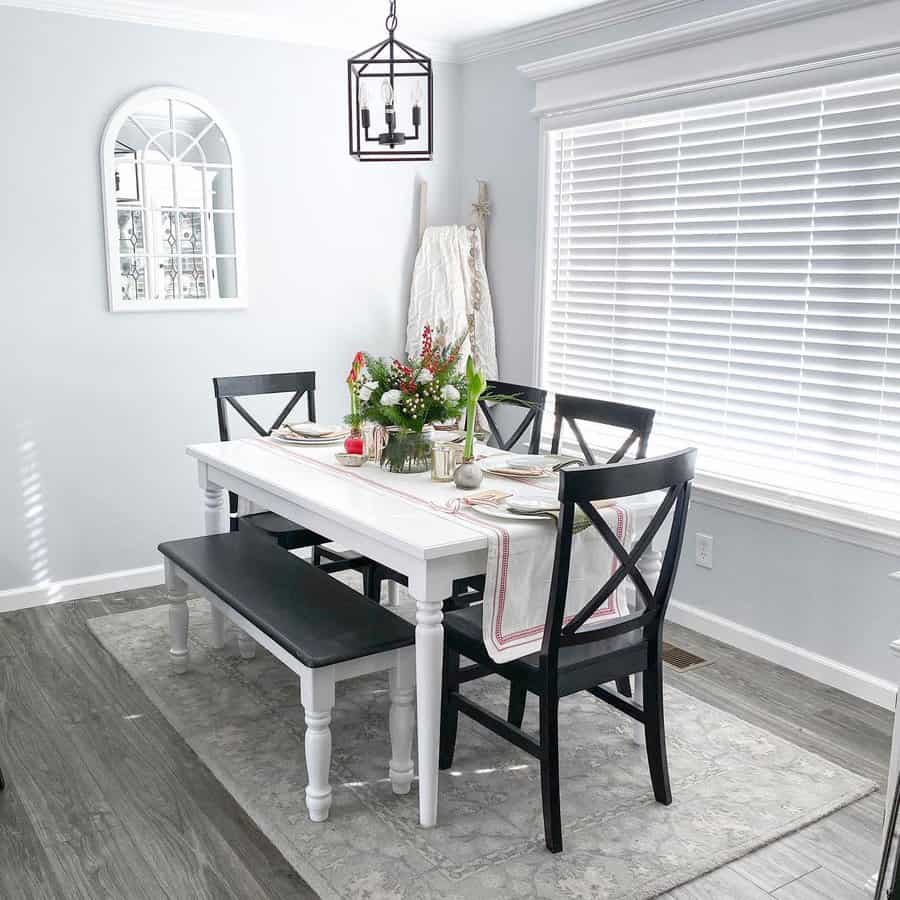 Dining area with a white table, black chairs, and a bench featuring a floral centerpiece on the table, large window with blinds in the background