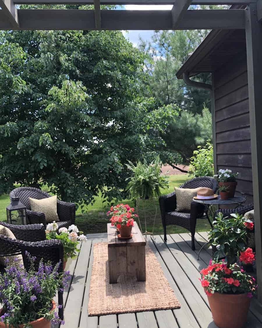 A cozy patio with wicker chairs, a wooden table, potted plants and flowers, surrounded by greenery under a pergola