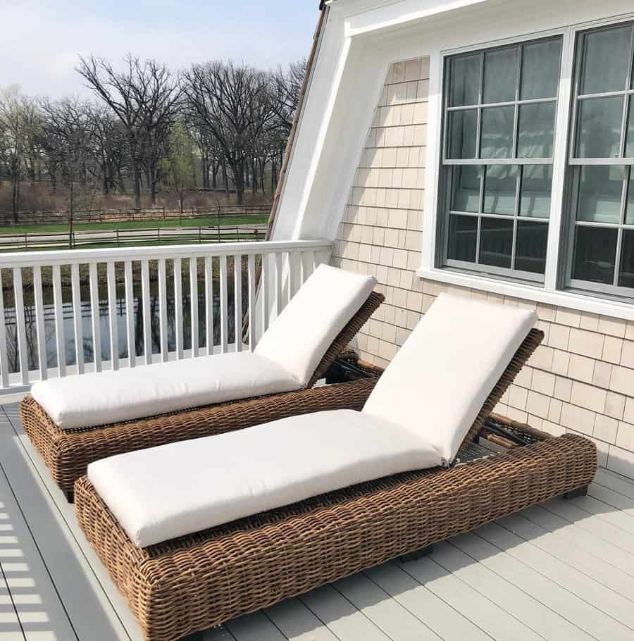 Two wicker chaise lounges with white cushions on a balcony with white railings overlooking a grassy area and trees