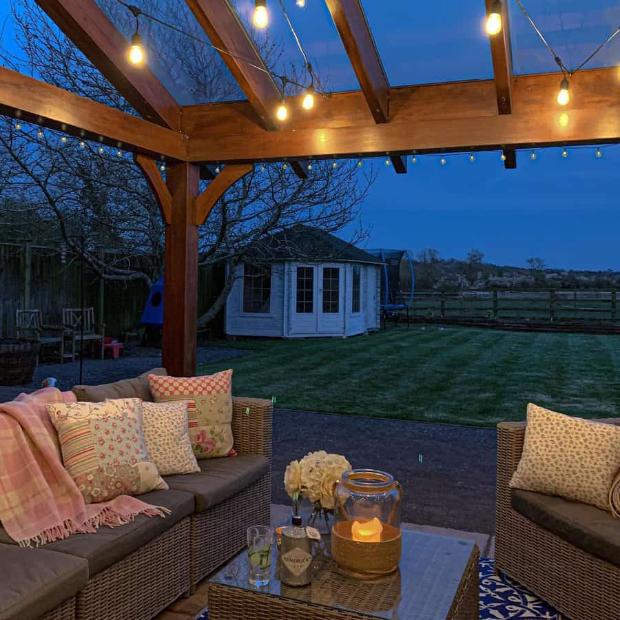 Cozy patio with wicker seating, colorful cushions, and string lights. Candle and flowers on the table. View of a garden and small shed