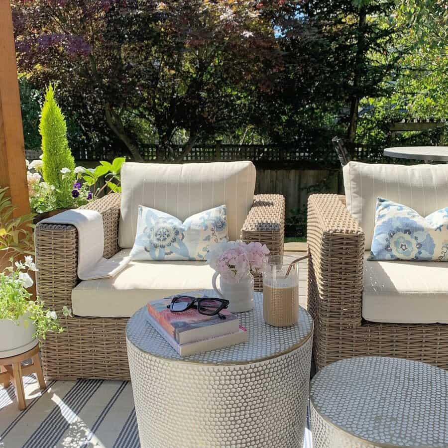 Outdoor patio with two wicker chairs, floral cushions, a round table with a book, sunglasses, a drink, and a flower vase surrounded by lush greenery