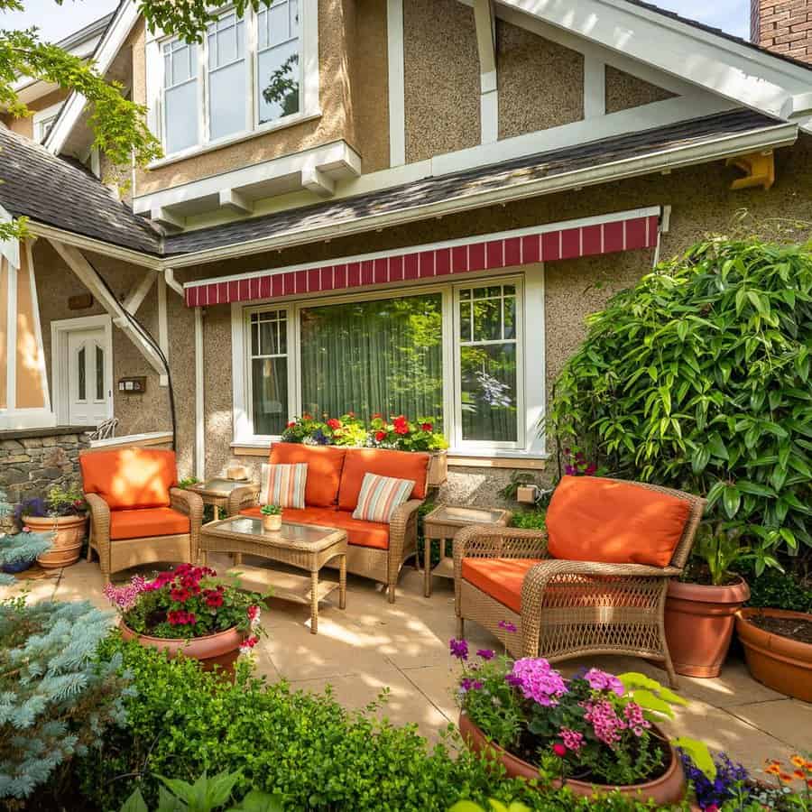 A cozy patio with wicker furniture and orange cushions, surrounded by lush greenery and colorful flowers under a striped red awning