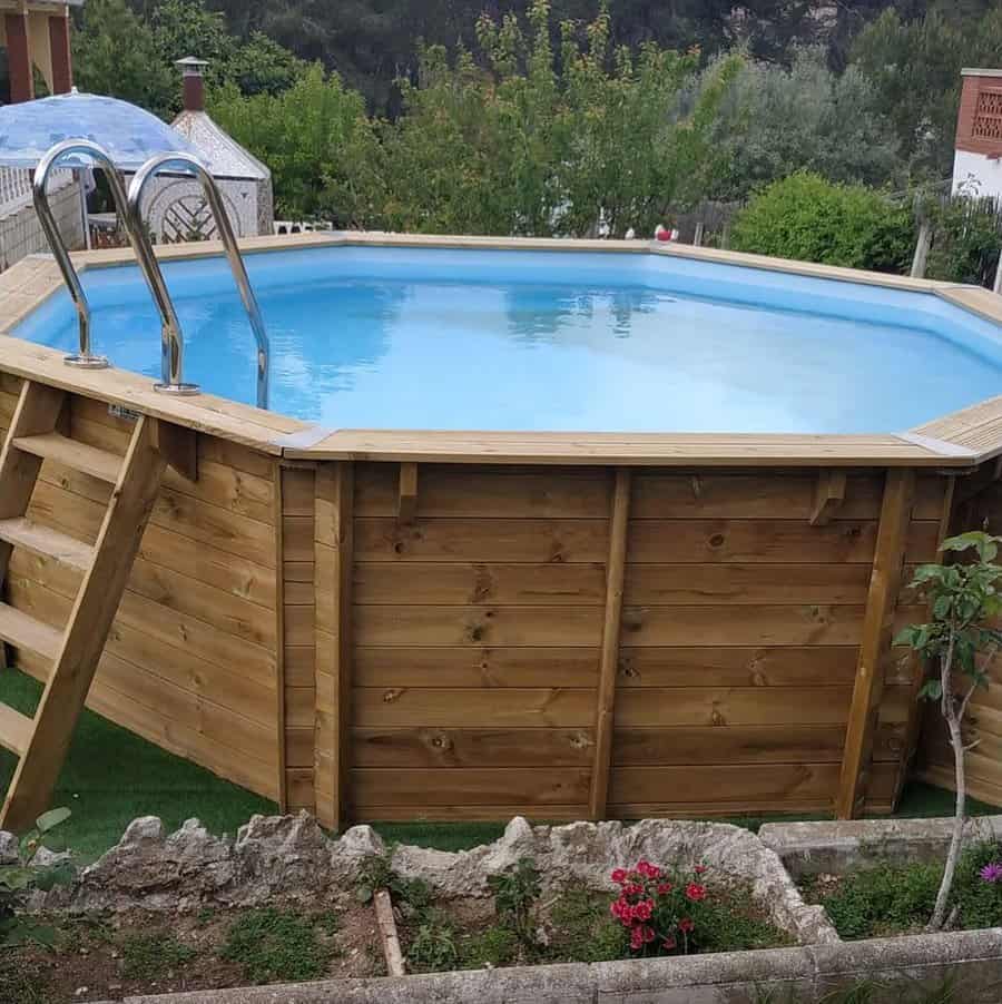 Octagonal above-ground pool with wooden panels, ladder, and metal handrails in a garden setting