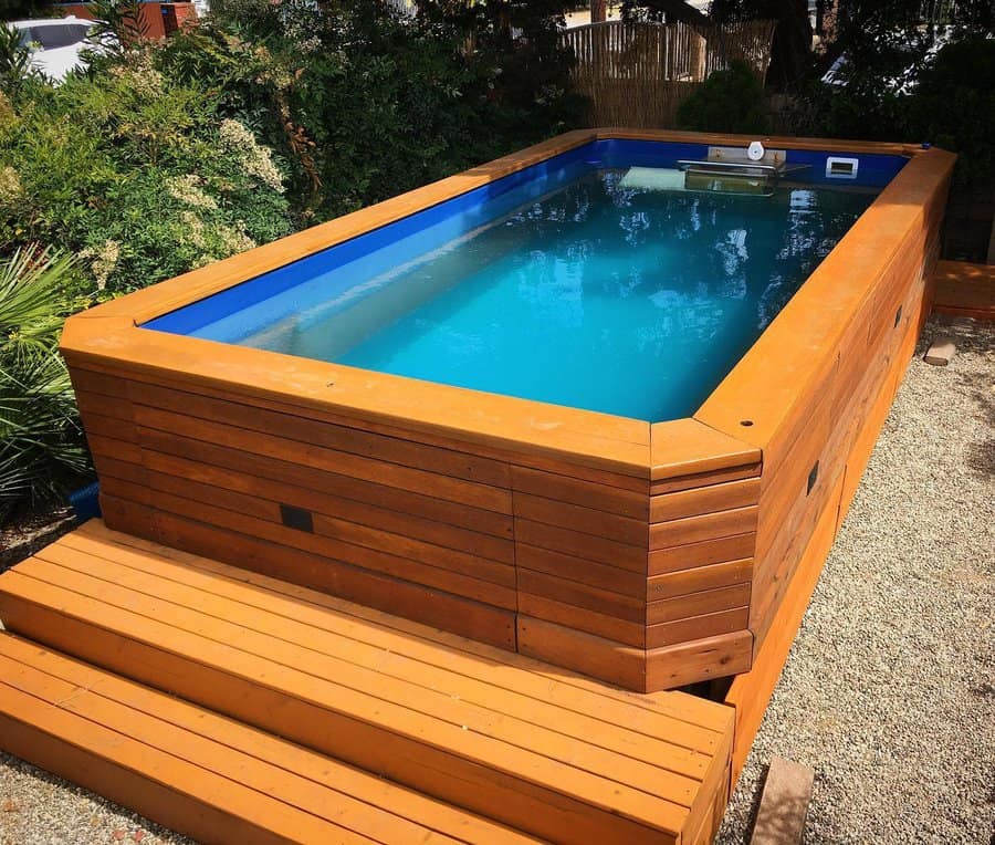 A small above-ground pool with wooden siding and steps, surrounded by plants and gravel