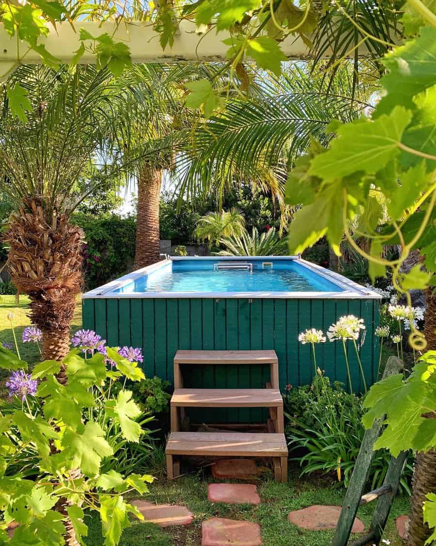 Above-ground pool in a lush garden setting with palm trees, flowers, and wooden steps leading to the water