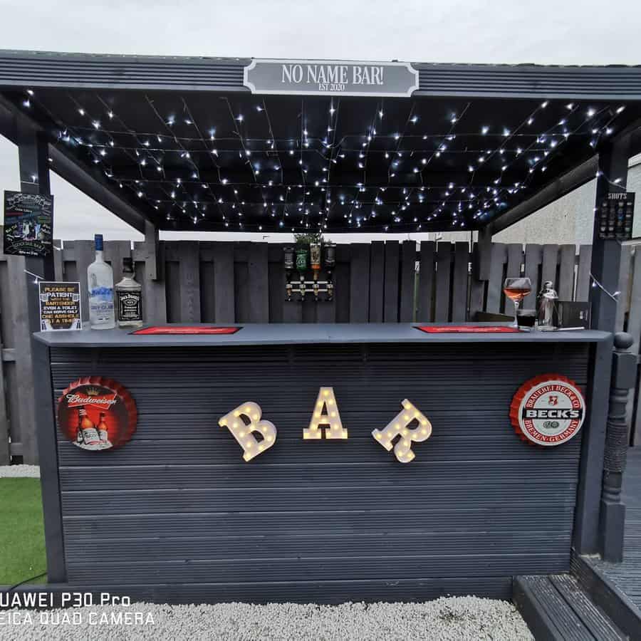 Outdoor bar setup with a wooden roof decorated with string lights, a counter labeled "BAR," and drink bottles on the shelves