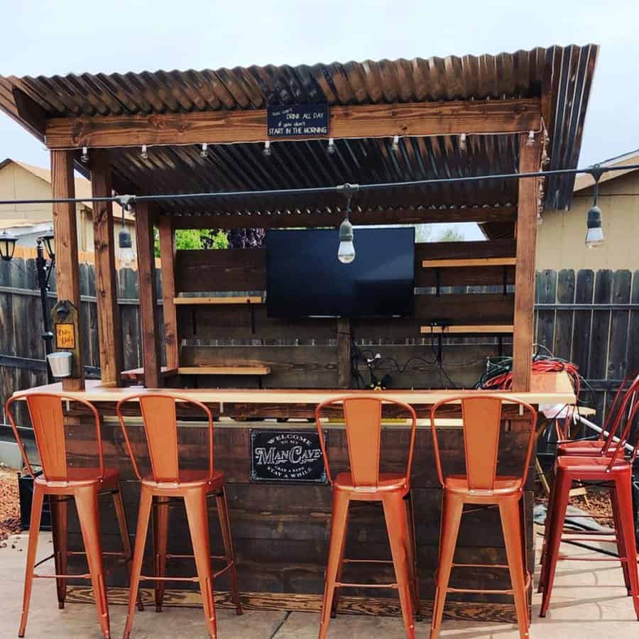 Outdoor wooden bar with a corrugated metal roof, four red metal stools, a wall-mounted TV, string lights, and "Man Cave" sign