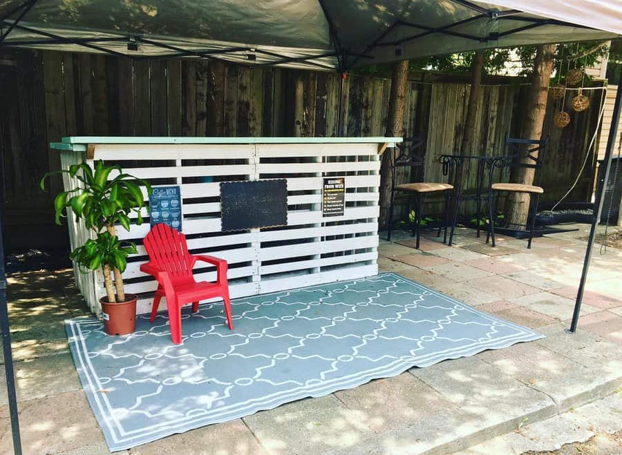 Outdoor setup with a white pallet bar, red chair, potted plant, and rug under a canopy; two bar stools in the background