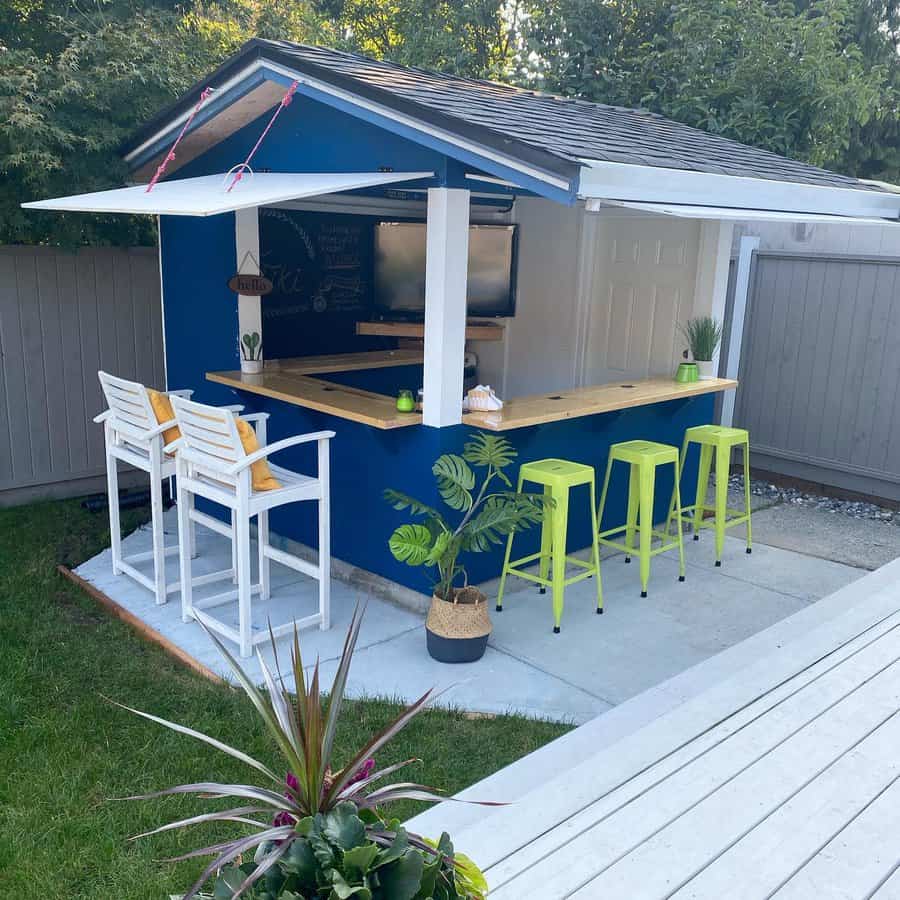 Backyard bar setup with blue walls, a wooden counter, white bar stools, and green stools, featuring plants and a TV visible