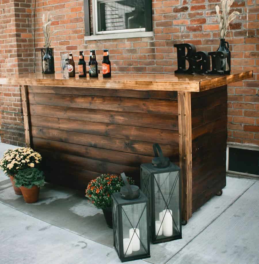 Wooden outdoor bar with beer bottles, "Bar" sign, two lanterns, and flowers, set against a brick wall