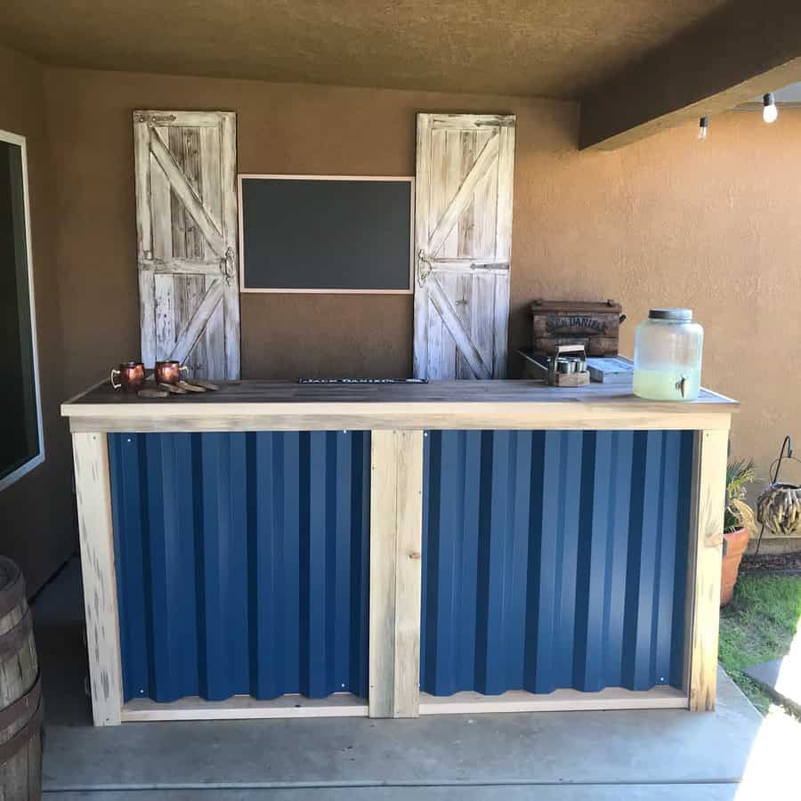 Outdoor bar with a rustic design, featuring blue corrugated metal panels, a wooden counter, and a chalkboard with barn-style shutters