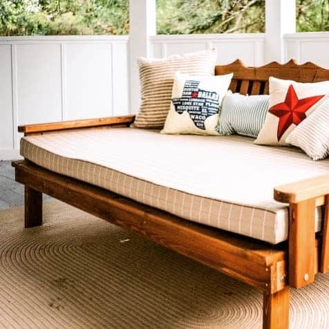 Rustic wooden daybed with a neutral cushion and patriotic-themed pillows, creating a cozy and inviting porch seating area