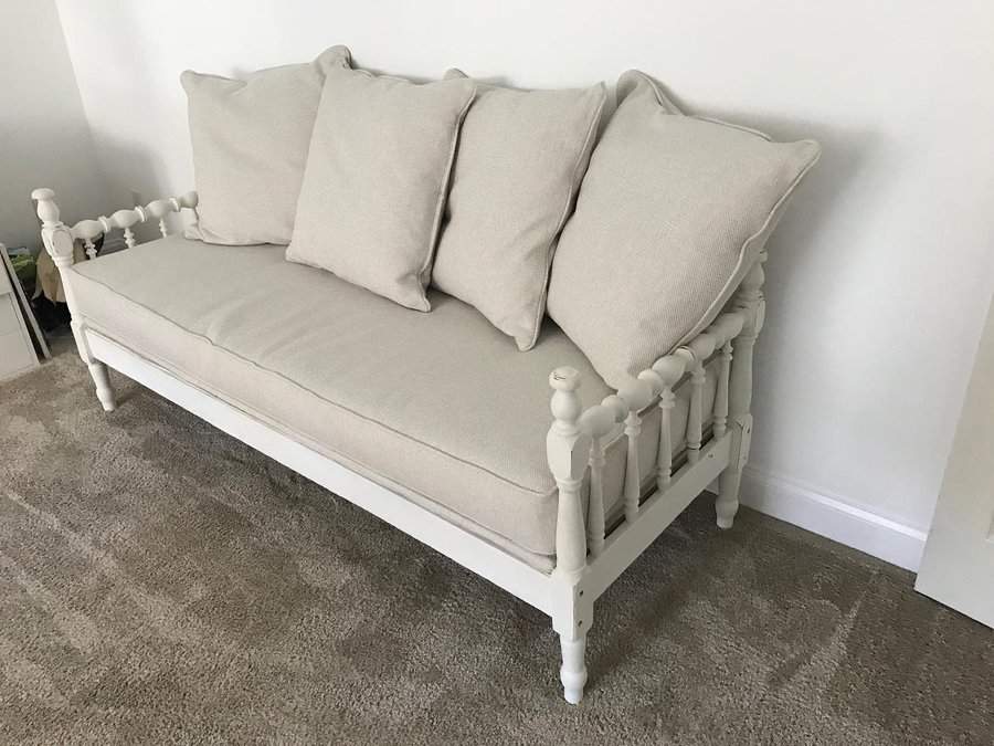 White wooden daybed with beige cushions against a white wall on a carpeted floor