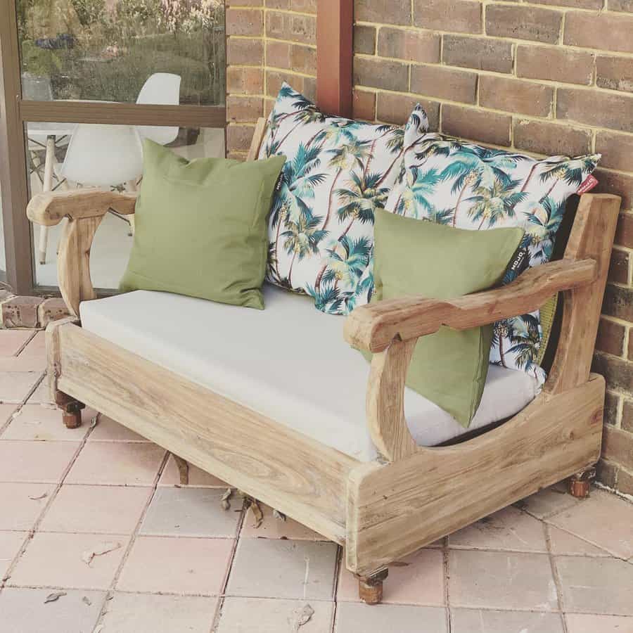 Wooden loveseat with tropical and green pillows on a patio with brick wall and glass window in the background