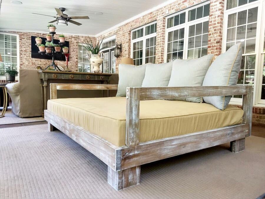 Wooden daybed with beige cushions on a sunlit porch, brick walls, large windows, ceiling fan, and potted plants in the background