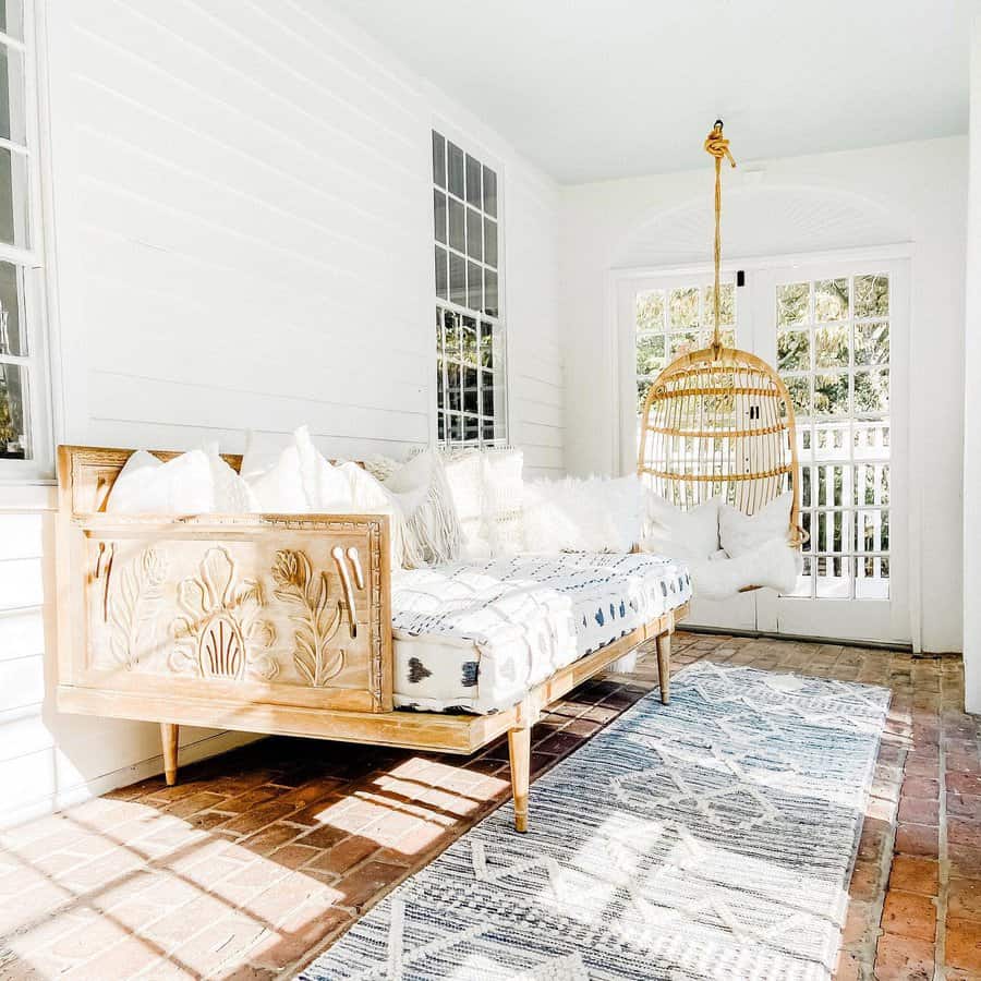 Boho-style carved wooden daybed with soft neutral cushions, patterned textiles, and a hanging rattan chair, creating a serene sunroom retreat