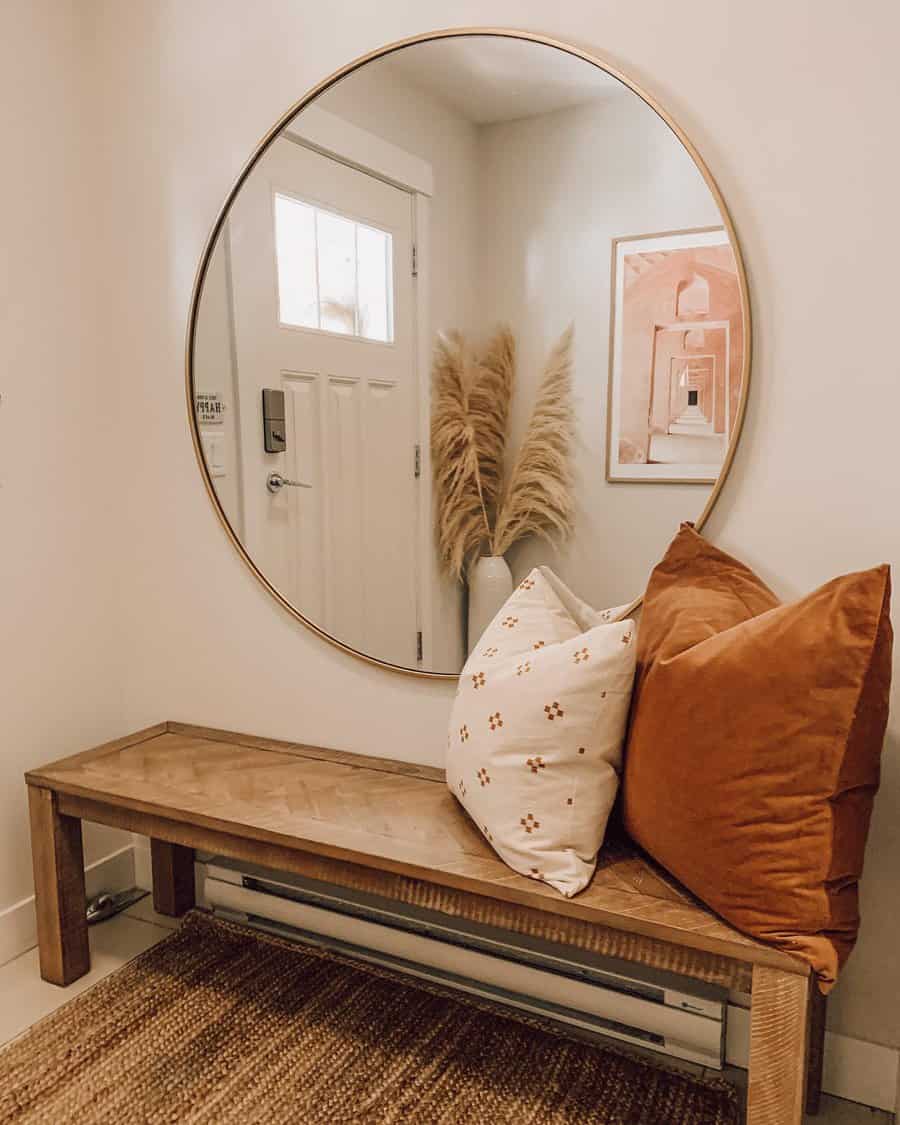 Entryway with large round mirror, wooden bench, orange and white pillows, vase with pampas grass, and art on the wall