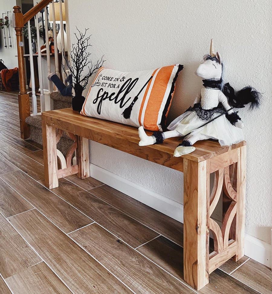 Wooden bench with Halloween decor: a “sit for a spell” pillow and two plush unicorns in costumes, brown tiled floor, staircase in background