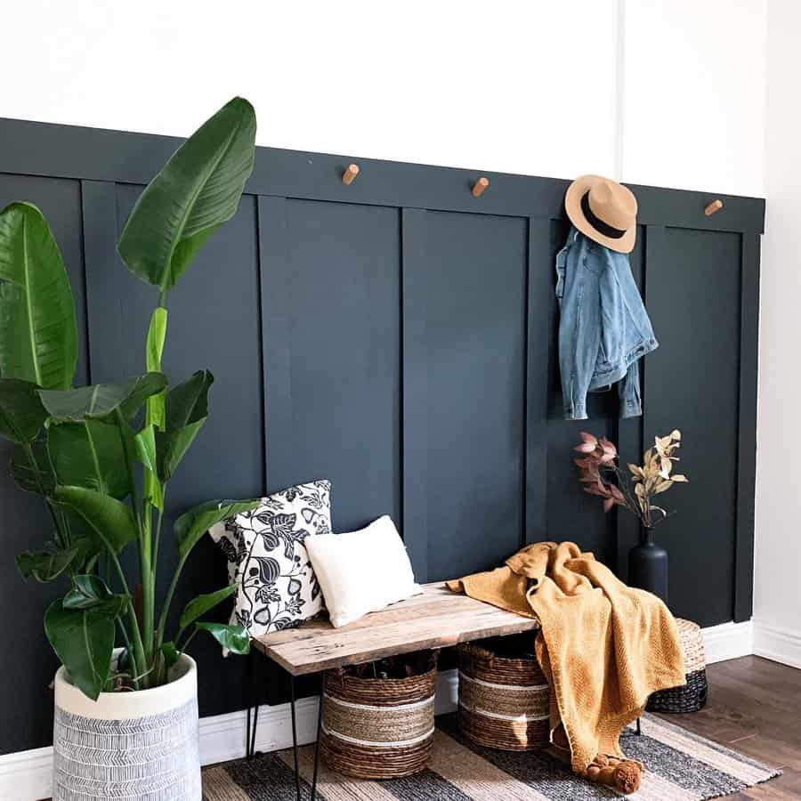 Cozy interior with dark green panel wall, wooden bench, pillows, large plant, and decor including a hat, jacket, and blanket