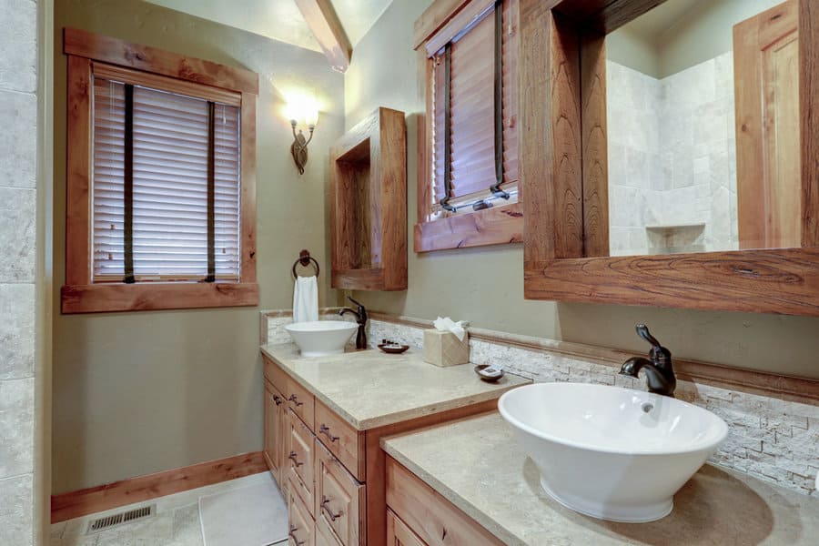 Rustic bathroom with wooden accents, two vessel sinks on a beige counter, and decorative mirrors. Window with blinds and wall light