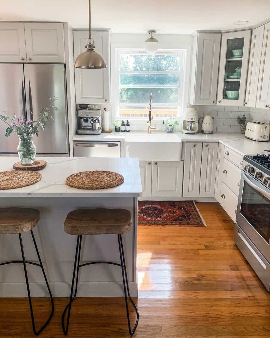 Warm hardwood flooring adds natural charm to this bright farmhouse kitchen, complementing white cabinetry, marble countertops, and rustic bar stools