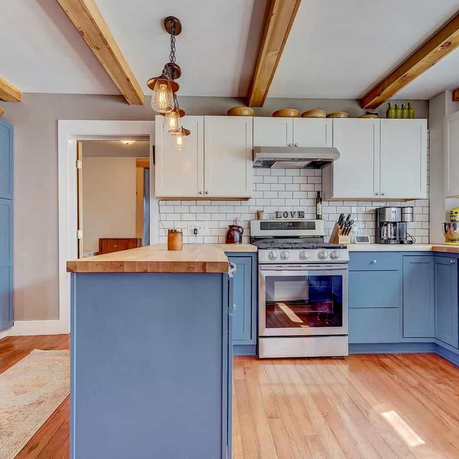 Warm hardwood flooring enhances this cozy farmhouse kitchen, complementing blue cabinetry, butcher block countertops, and exposed wooden ceiling beams