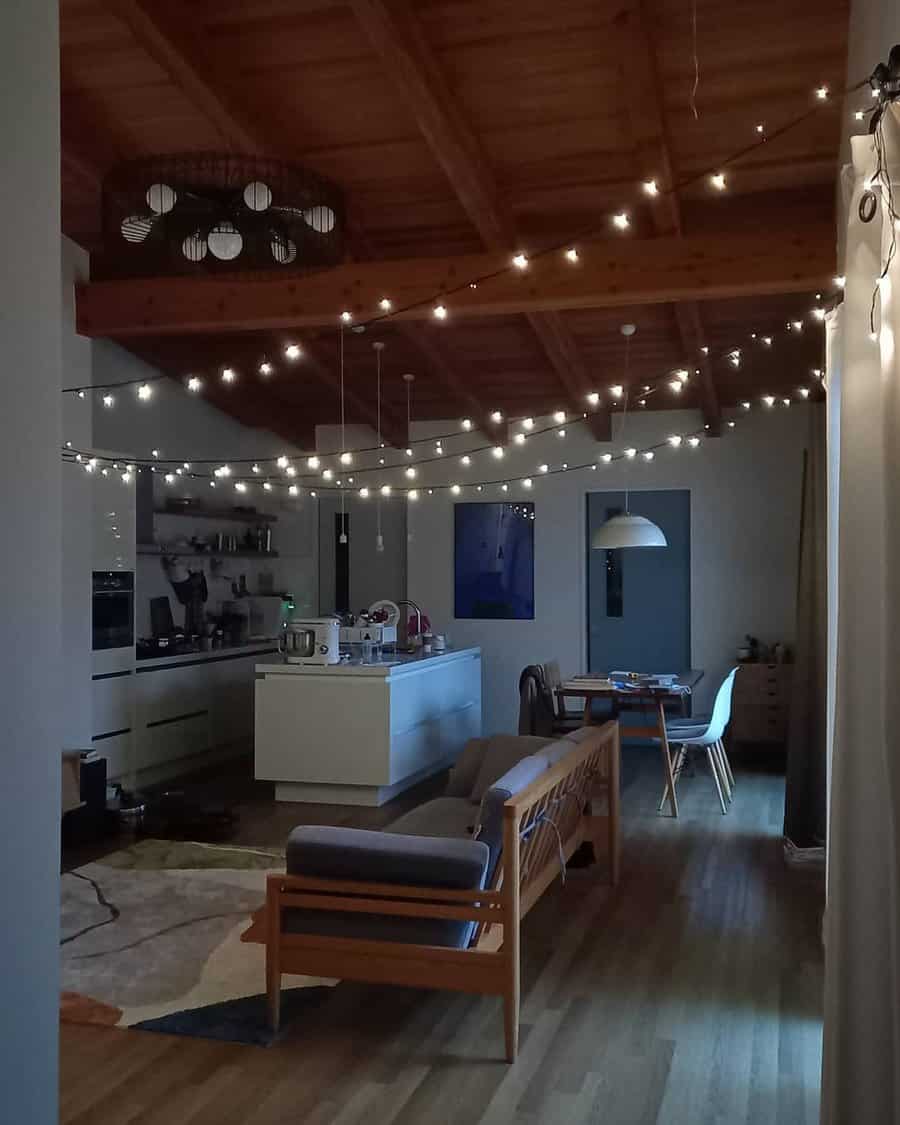 Small living room with string lights, wooden ceiling, modern kitchen, couch, and dining area creating a warm, inviting atmosphere