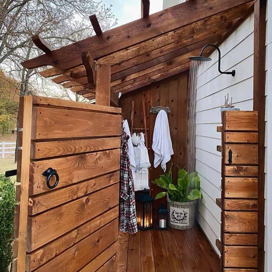 Outdoor wooden shower with a rustic design, featuring a rain showerhead, towels, potted plant, and lanterns on a wooden deck