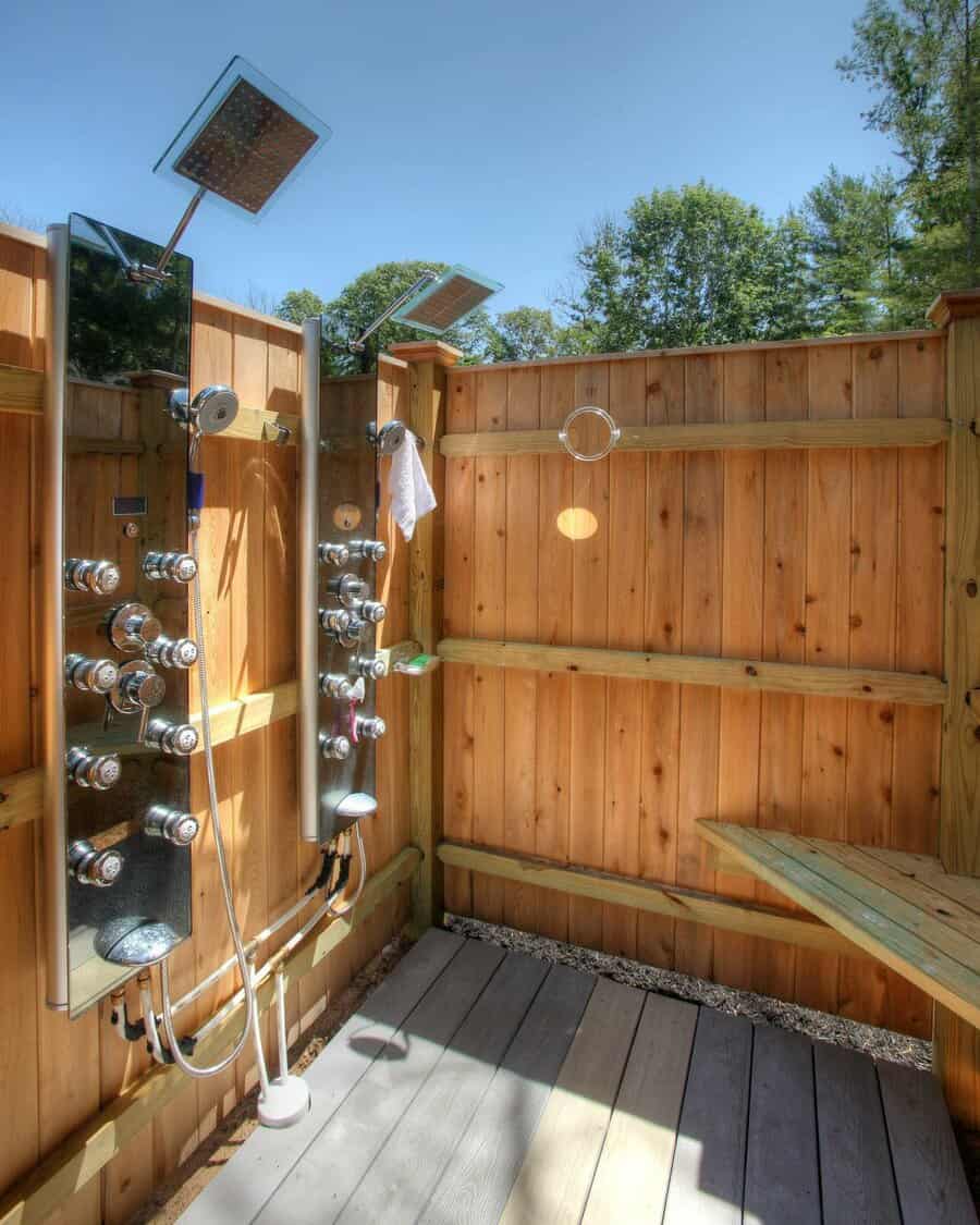 Outdoor shower with wooden walls, multiple shower heads, a towel, and a small bench on a bright and sunny day