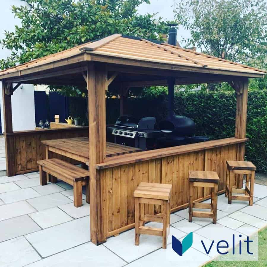 Wooden outdoor gazebo with a seating area and barbecue grill, surrounded by greenery